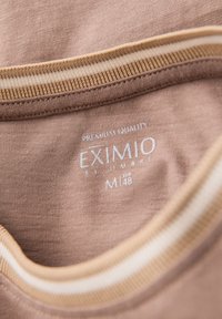 Braunes T-Shirt mit weichem Material, ausgestattet mit einem gerippten Kragen in Beige mit weißen Streifen und einem aufgedruckten Etikett, das Marken- und Größeninformationen zeigt.