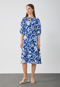 Robe à motif floral dans un bold bleu et blanc, dotée d'un décolleté en trou de serrure et d'une taille froncée. Mi-longue avec des manches bouffantes, associée à des talons légers.
