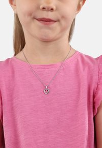 Collana d'argento con un ciondolo a forma di cuore, caratterizzato da un cuore interno rosa e un bordo di pietre trasparenti, indossata sopra una camicia di cotone rosa.