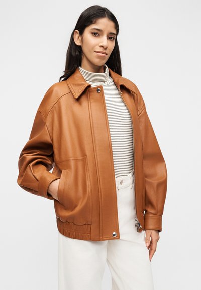 Γυναίκα που φορά καφέ δερμάτινο μπουφάν πάνω από ριγέ πουλόβερ με ψηλό λαιμό και άσπρο παντελόνι, στέκεται με το ένα χέρι στην τσέπη του μπουφάν μπροστά σε απλό φόντο.