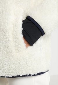 Vit, fluffig fleecejacka med en mjuk textur, med manchetter och nederkant i marinblått tyg. Inkluderar en ficka på sidan.