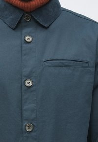 Sötét türkizszínű ing kordbársony gallérral, elöl hat gombbal és egy kis patentos zsebfedővel a bal oldalon.