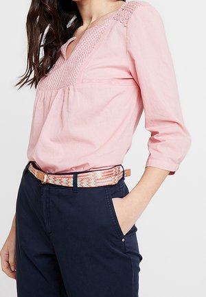 Vrouw draagt een roze textuur V-hals blouse en marineblauwe broek met een patroon geweven riem, hand in de zak, tegen een effen achtergrond.