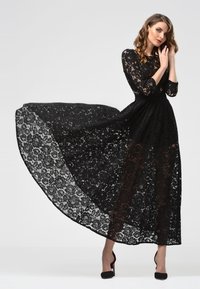 Schwarzes Spitzenkleid mit langen Ärmeln, fließender Silhouette, durchscheinendem unteren Teil, floralem Muster, tailliertem Bund, kombiniert mit schwarzen High Heels.