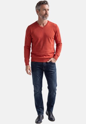 Mann mit salz-und-pfefferfarbenem Haar, der einen roten V-Ausschnitt-Pullover, dunkelblaue Jeans und schwarze Schuhe trägt, mit einer Hand in der Tasche vor weißem Hintergrund steht.