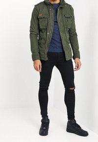 Olivgrön bomulljacka med dragkedja, fyra framfickor och axelklaffar, kombinerad med svarta rivna slim-fit jeans och svarta sneakers.