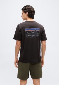 Νεαρός άνδρας με την πλάτη, φοράει μαύρο μπλουζάκι Patagonia με γραφικό βουνού και το κείμενο «Save Our Home Planet», συνδυασμένο με χακί σορτς.