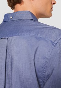 Blaue Jeanshemd mit Button-Down-Kragen, glatter Textur und einer Faltendetail auf dem Rücken. Subtile Nähte akzentuieren das Design.