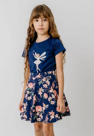 Girl with long wavy hair wearing a navy blue dress featuring a fairy print and floral patterned skirt, standing against a plain light background.