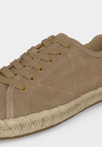 Gros plan d'une basket en suède beige avec des lacets assortis, une semelle intermédiaire en corde tressée et "GUESS" embossé sur la partie latérale.
