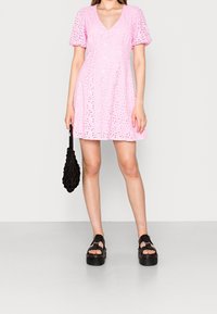 Žena ve světle růžových šatech s volnými rukávy, držící černou síťovanou tašku, v černých platformových sandálech, stojící na šedé podlaze proti bílé zdi.