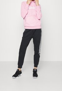 Różowa bluza z kapturem z białym logo, czarne zwężane spodnie dresowe z białymi paskami po bokach oraz czarne buty sportowe.