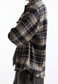 Personne portant une chemise en laine à carreaux noir, beige et gris avec un ourlet effiloché sur un pantalon noir, les bras croisés, vue de côté.