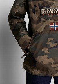 Kurtka w kamuflażu w odcieniach zieleni i brązu, z zamykaną kieszenią, naszywką z logo i akcentem w postaci norweskiej flagi z przodu.
