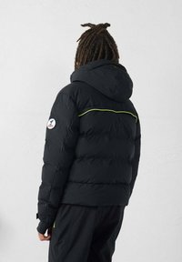 Zwarte gewatteerde jas met capuchon, voorzien van een horizontale gele bies en een logopatch op de linker mouw.