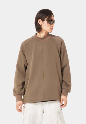 Sweatshirt marron avec un col haut, des manches raglan et un subtil motif de couture à l'avant. Le modèle porte des lunettes de soleil au design élégant.