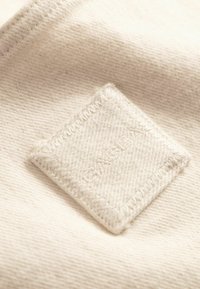 Nærbillede af et blødt beige stof med en diamantformet, syet lap, der viser det diskrete broderede ord "CLOSET".