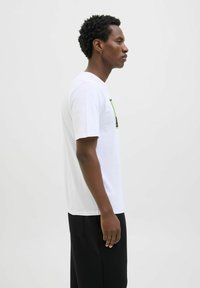 Weißes Baumwoll-T-Shirt mit einem grünen und schwarzen Grafikdruck auf der Vorderseite, kombiniert mit schwarzen Hosen, Modell steht im Profil.