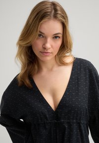 Zwarte blouse met een V-hals, voorzien van een subtiel geometrisch patroon. Zachte stof, losse pasvorm en geraffineerd detail bij de taille.