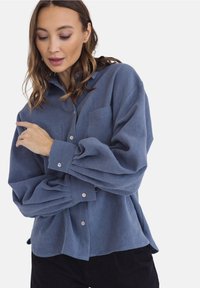 Niebieska koszula z guzikami, o miękkiej fakturze, z dwiema przednimi kieszeniami i zgromadzonymi rękawami. Posiada luźny krój i zaokrąglony dół.