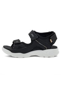 Svart sandal med en strukturerad design, utrustad med justerbara kardborreband och en kraftig vit sula. Tillverkad av andningsbara material med nätinslag.