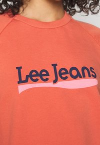 Korálový svetr s kulatým výstřihem, raglánovými rukávy a tmavě modrým logem "Lee Jeans" s růžovým akcentem pod textem. Hladký povrch.