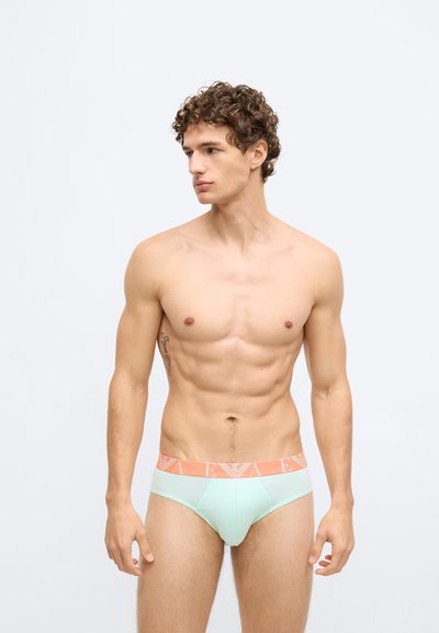 Cuecas de algodão verde claro com uma cintura laranja que apresenta um logótipo. O tecido parece suave com um design ajustado.
