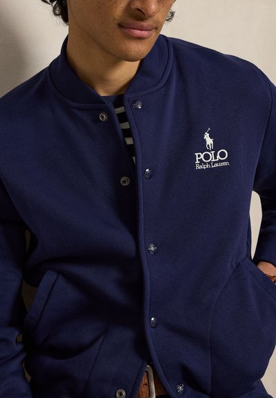 Άνδρας που φοράει ένα ναυτικό μπλε σακάκι Polo Ralph Lauren με κουμπιά και λευκό λογότυπο στο αριστερό στήθος, ριγέ πουκάμισο από κάτω, τα χέρια στις τσέπες.