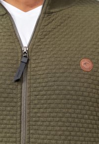 Homme portant une veste zippée texturée vert olive avec une fermeture éclair argentée et un petit patch rond marron avec logo sur la poitrine, par-dessus une chemise blanche.