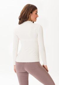 Top bianco a maniche lunghe con scollo a scallop e design aderente, abbinato a leggings color malva, che mostrano una superficie liscia.