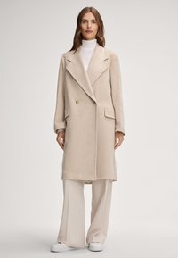 Beige dubbele jas met dubbele rij knopen van zachte stof, met grote revers, voorzakken en gouden knopen, gedragen over een witte coltrui en lichte broek.