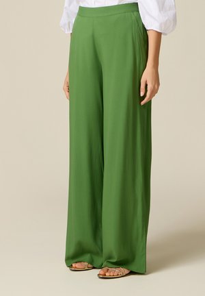 Mujer que lleva pantalones verdes de talle alto y pata ancha con bolsillos laterales y sandalias doradas de tiras, combinados con una blusa blanca.