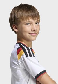 Ungdoms-fotbollströja med vitt tyg, svarta och gula accenter, texturerad yta och horisontella svarta ränder på axlarna.