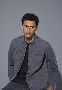 Dunkelgrauendes Hemd aus Denim mit zwei Brusttaschen und Druckknöpfen. Glatte Textur, lange Ärmel und eine leicht lockere Passform, kombiniert mit einem schwarzen Oberteil.