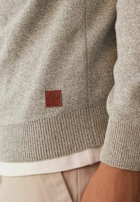 Šviesiai pilkas megztinis, pagamintas iš minkštos medžiagos, su kutura apatinėje dalyje ir rankogaliuose, ant kurio yra mažas rudas odinis ženklelis su logotipu.