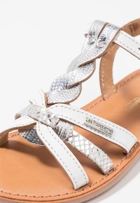 Bílé sandály s pletenými stříbrnými akcenty, hladkými koženými popruhy a hnědou stélkou s metalickou logem.