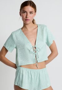 Top corto de un verde menta claro con escote en V, mangas cortas y un detalle de lazo en la parte delantera, combinado con pantalones cortos a juego en un material similar.