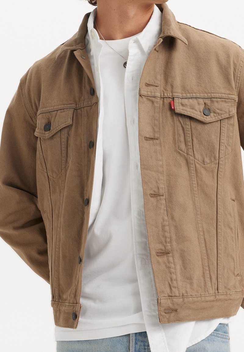 Homme portant une veste marron clair boutonnée par-dessus une chemise blanche déboutonnée et un t-shirt blanc avec un jean bleu clair.