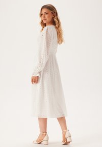 Weißes, langärmeliges Kleid mit Lochmuster, tailliertem Bund und halbtransparentem Stoff, kombiniert mit hellen Blockabsatzsandalen.