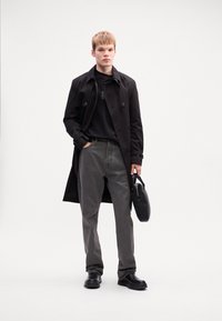 Black trench coat over a black shirt, gray pants, and black shoes. The model holds a black bag and stands against a light background.
