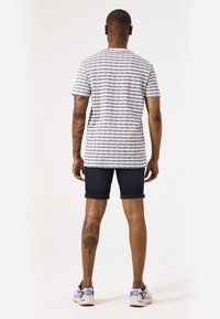 Camisa de manga corta para hombre con rayas horizontales en navy y blanco, combinada con pantalones cortos de mezclilla oscuros. Tatuajes visibles en las piernas y zapatillas deportivas.