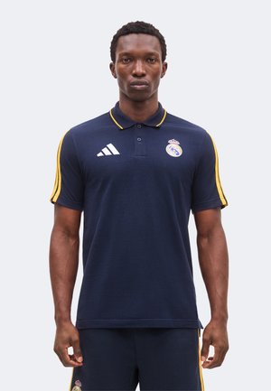 Homme portant un polo bleu marine du Real Madrid avec des bandes jaunes sur les manches et le logo Adidas, debout devant un fond uni.