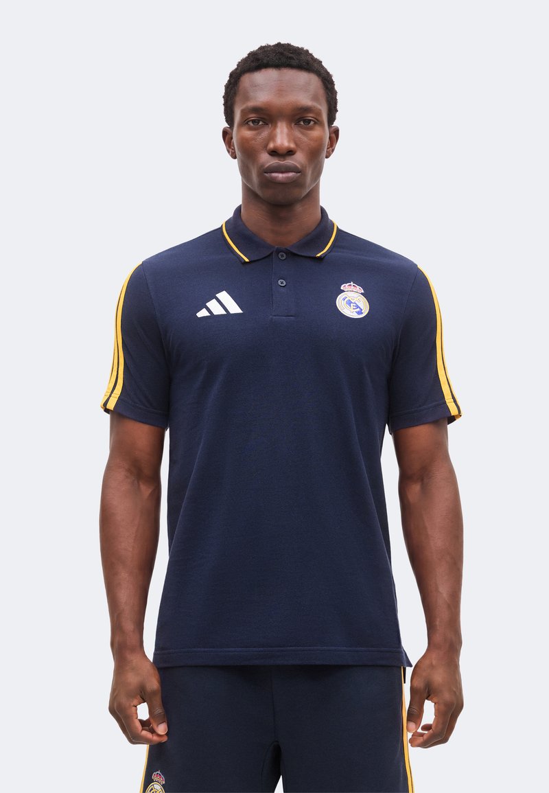Homme portant un polo bleu marine du Real Madrid avec des bandes jaunes sur les manches et le logo Adidas, debout devant un fond uni.