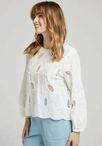 Blouse blanche brodée avec des motifs floraux, ourlet festonné et zones transparentes. Présente des manches longues et un col rond.
