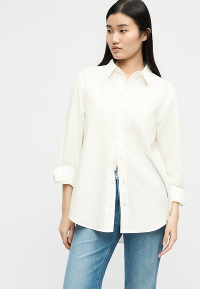 Νεαρή γυναίκα με μακριά σκούρα μαλλιά, φορώντας άνετο λευκό πουκάμισο με κουμπιά και γυρισμένα μανίκια, και ανοιχτόχρωμα μπλε τζιν, στέκεται μπροστά σε απλό φόντο.