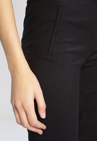Svarta byxor gjorda av ett slätt, elastiskt tyg med slim fit och en sidficka. Handen vilar på låret, vilket framhäver textur och design.