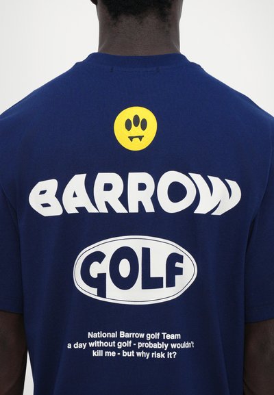 Tummansininen t-paita, jossa on suuret valkoiset tekstit "BARROW GOLF", ja keltainen hymyilevä naama, jolla on kulmahampaat. Mukana myös lisäteksti valkoisena alla.