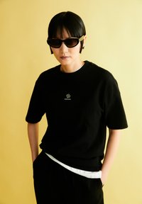 Zwarte T-shirt met korte mouwen met een bloemenlogo op de borst, gecombineerd met zwarte broek en zonnebril, tegen een felgele achtergrond.