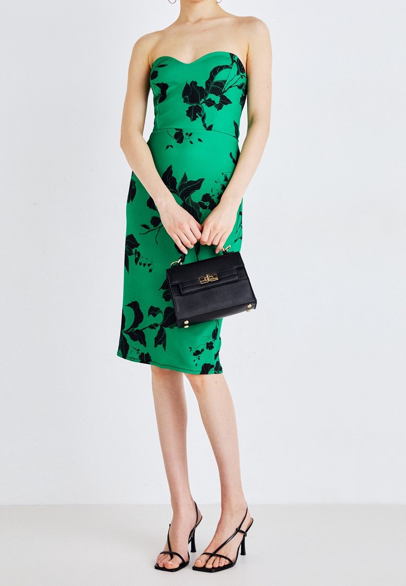 Strapless grøn midi-kjole med sort blomstermønster, tætsiddende design. Modellen holder en lille sort håndtaske med gyldne accenter og har sorte strop-sandaler på.