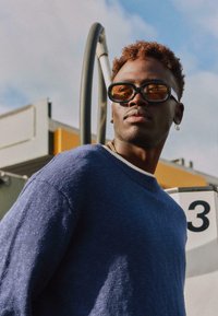 Jeune homme portant des lunettes de soleil foncées, un pull bleu et une boucle d'oreille unique, debout près d'une pompe à essence numéro 3 sous un ciel partiellement nuageux.
